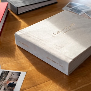 May include: A white velvet wedding album with gold lettering on the cover. The text reads "Our Wedding" and "Jonathan & Samantha" with the date "September 17, 2015".