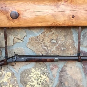 May include: A long, dark brown rifle with a metal barrel and a wooden stock is hanging on a wall. The rifle is secured with two leather straps. The wall is made of stone and has a wooden beam above it.