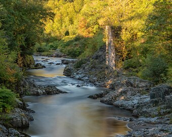 Puede incluir: Un río fluye a través de una zona rocosa y arbolada. El agua es lisa y los árboles son verdes y amarillos. El sol brilla sobre el agua, creando un brillo dorado.