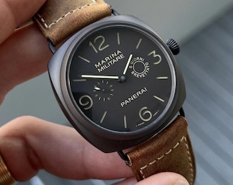 May include: A Panerai Marina Militare watch with a matte black cushion-shaped case and a black dial featuring luminous Arabic numerals at 3, 6, 9, and 12 o'clock. The watch includes a brown suede leather strap with white contrast stitching.