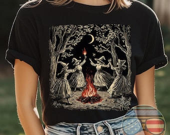 May include: Black t-shirt with a graphic print of four witches dancing around a bonfire in a forest. The witches are wearing long dresses and pointy hats. The moon is visible in the sky.