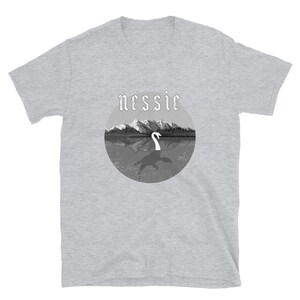May include: A gray t-shirt with a black and white graphic of a lake with mountains in the background. The graphic has the word "nessie" in a gothic font above the lake.