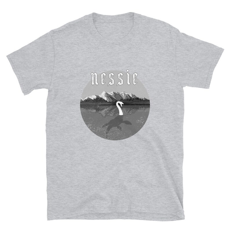 May include: A gray t-shirt with a black and white graphic of a lake with mountains in the background. The graphic has the word "nessie" in a gothic font above the lake.