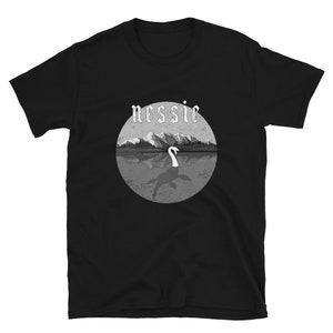 May include: Black t-shirt with a graphic of a gray and white Loch Ness Monster in a lake with mountains in the background. The text "nessie" is above the graphic in a white gothic font.