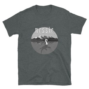 May include: A dark heather grey t-shirt with a black and white illustration of the Loch Ness Monster, Nessie, in a circle with the word "nessie" in a gothic font above the illustration.