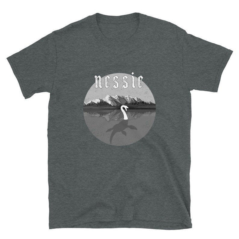 May include: A dark heather grey t-shirt with a black and white illustration of the Loch Ness Monster, Nessie, in a circle with the word "nessie" in a gothic font above the illustration.