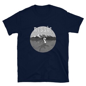 May include: A navy blue t-shirt with a graphic of a lake monster, Nessie, in a lake with mountains in the background. The text "nessie" is in a gothic font.