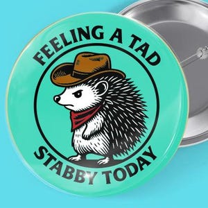 May include: A 2.25-inch button pin with a teal background featuring a cartoon hedgehog wearing a cowboy hat and a red bandana. The text reads "Feeling a tad stabby today." A silver pin back is visible.