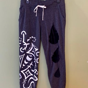 May include: Dark gray sweatpants with a white drawstring and pockets. The left leg features a white graphic design, while the right leg has three black velvet teardrop shapes. The pants are hanging on a black hanger.