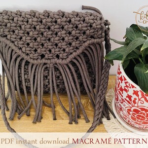 Puede incluir: Un bolso de macram&eacute; marr&oacute;n con flecos. El bolso tiene una correa larga y est&aacute; hecho con un cord&oacute;n grueso. El bolso est&aacute; sobre una superficie de madera con una maceta de patr&oacute;n blanco y rojo y una planta verde en el fondo. El texto "PDF instant download MACRAM&Eacute; PATTERN" est&aacute; en la parte inferior de la imagen.