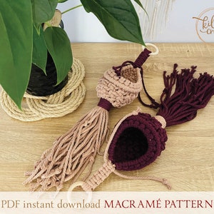 Peut inclure: Deux suspensions de plantes en macram&eacute; dans un sch&eacute;ma de couleurs rose et bordeaux. Les suspensions sont faites avec du cordon de coton et ont un pompon en bas. Les suspensions sont suspendues &agrave; une surface en bois. Le texte "PDF instant download MACRAME PATTERN" est en bas de l'image.