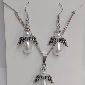 May include: A silver necklace and earrings set featuring angel pendants with white pearl accents. The necklace has a silver chain and a pendant with a larger pearl.