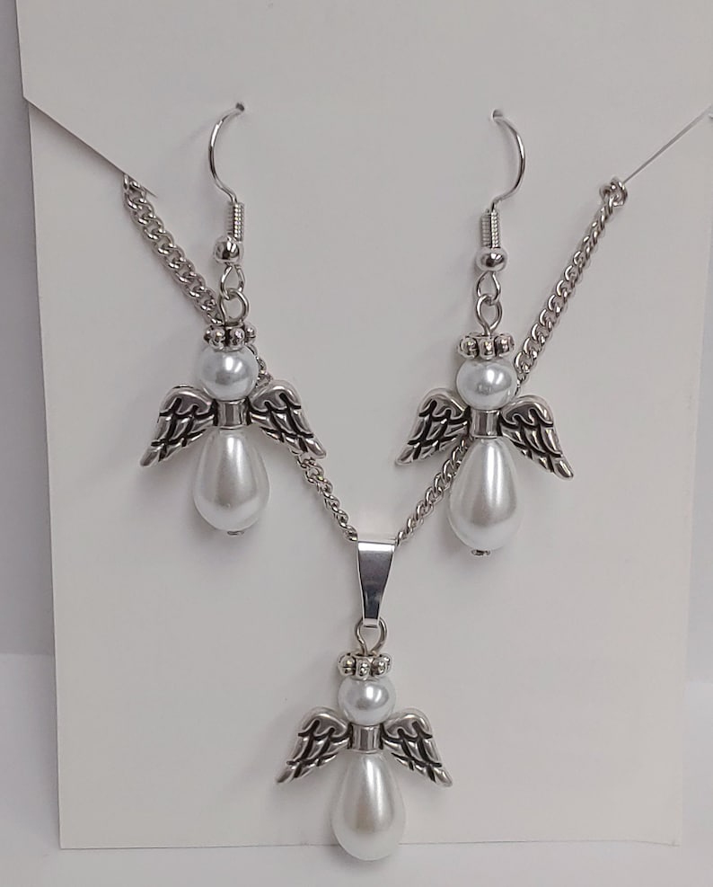 May include: A silver necklace and earrings set featuring angel pendants with white pearl accents. The necklace has a silver chain and a pendant with a larger pearl.