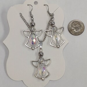 May include: A silver angel necklace and earrings set with iridescent crystal accents. The set is displayed on a white card with a silver chain and a silver angel pendant.