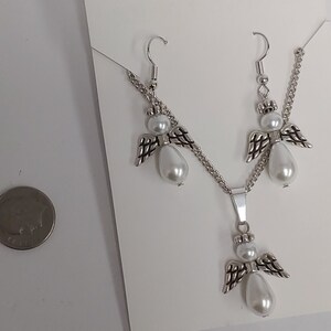 May include: A silver necklace and earrings set featuring angel charms with white pearl accents. The necklace has a silver chain and a pendant with an angel charm. The earrings are silver with angel charms and white pearl accents.