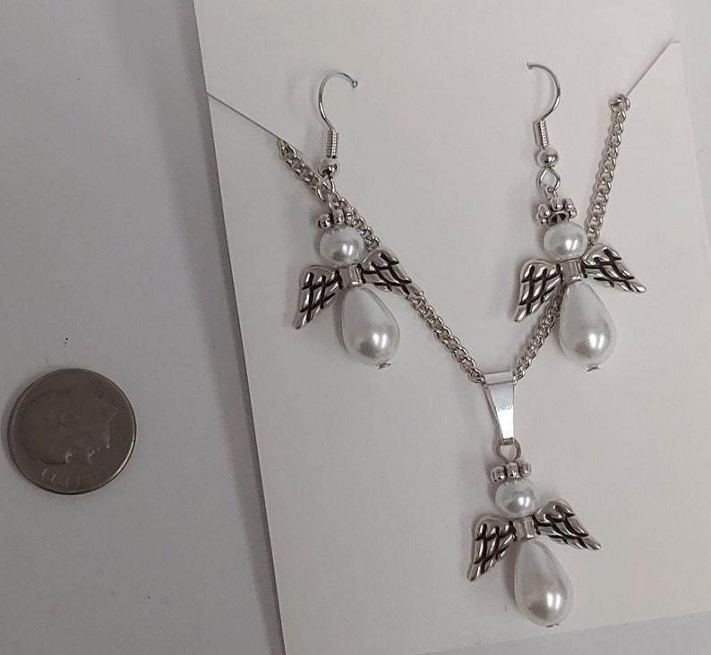 May include: A silver necklace and earrings set featuring angel charms with white pearl accents. The necklace has a silver chain and a pendant with an angel charm. The earrings are silver with angel charms and white pearl accents.