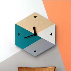 May include: Geometric wooden wall clock with teal, gray, white, and natural wood sections. The clock has black hands and black triangle markers. The clock is mounted on a wall with a gray and orange background. The clock is a hexagon shape.