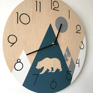 Close-up of a round wooden clock with a geometric mountain scene, bear and deer silhouettes, and a grey sun. Features clean black Arabic numerals and minimalist hands, highlighting the natural wood grain