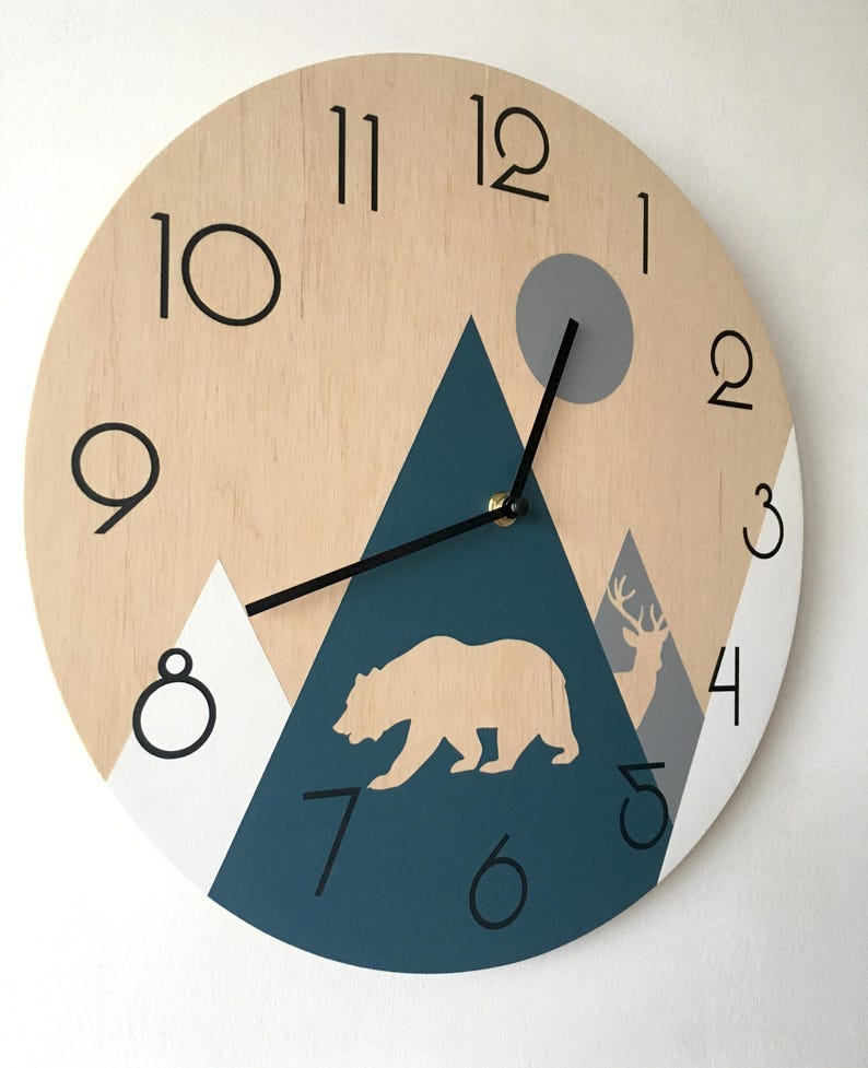 Close-up of a round wooden clock with a geometric mountain scene, bear and deer silhouettes, and a grey sun. Features clean black Arabic numerals and minimalist hands, highlighting the natural wood grain