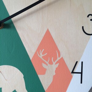 Close-up of a round wooden clock with a geometric mountain scene, deer silhouette. Features clean black Arabic numerals and minimalist hands, highlighting the natural wood grain