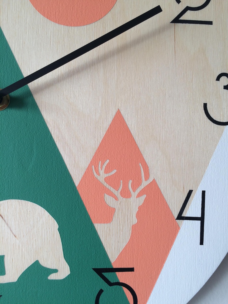 Close-up of a round wooden clock with a geometric mountain scene, deer silhouette. Features clean black Arabic numerals and minimalist hands, highlighting the natural wood grain