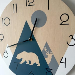 Side angle close-up of a wooden wall clock featuring a mountain landscape design. The image shows the natural wood grain texture with painted navy blue and grey mountains, a bear silhouette, and a deer with antlers. Minimalist black numerals and slim black hands are clearly visible against the light wood surface