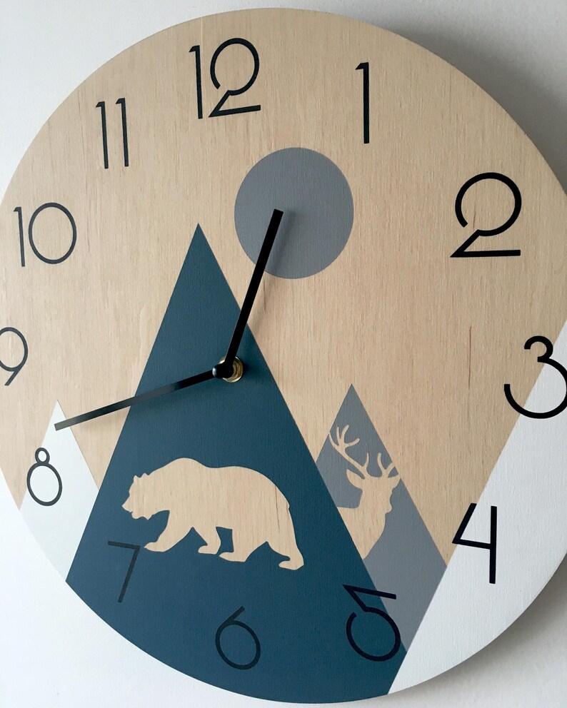 Side angle close-up of a wooden wall clock featuring a mountain landscape design. The image shows the natural wood grain texture with painted navy blue and grey mountains, a bear silhouette, and a deer with antlers. Minimalist black numerals and slim black hands are clearly visible against the light wood surface