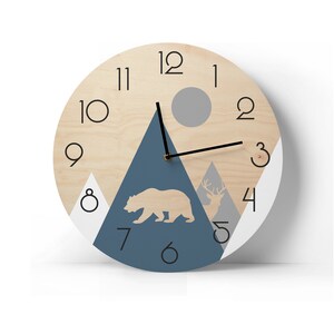 Round wooden wall clock with a geometric mountain landscape, bear and deer silhouettes. Modern rustic home decor with large black numbers and minimalist hands, shown against a bright white wall