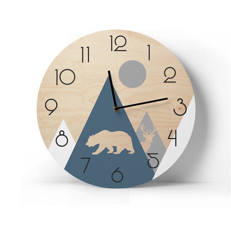 Round wooden wall clock with a geometric mountain landscape, bear and deer silhouettes. Modern rustic home decor with large black numbers and minimalist hands, shown against a bright white wall
