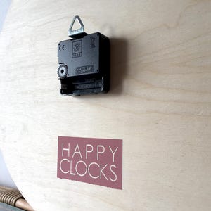 A close-up of the back of a round wooden clock with a black clock mechanism and a red label that says "HAPPY CLOCKS".