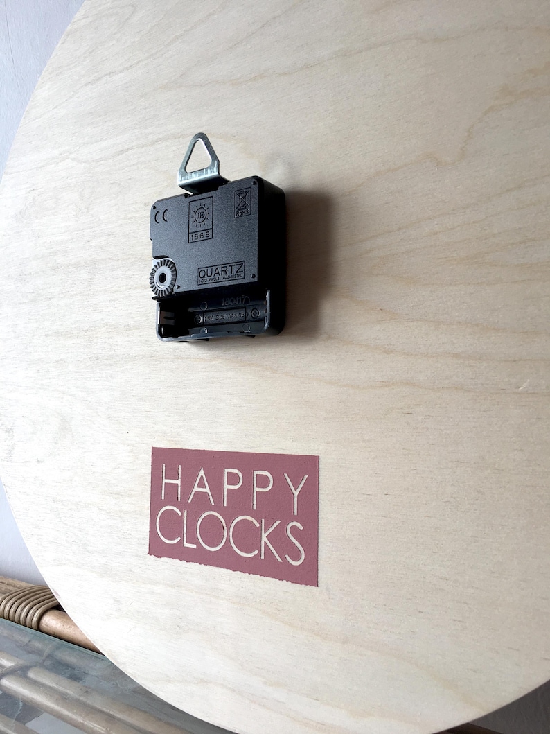 A close-up of the back of a round wooden clock with a black clock mechanism and a red label that says "HAPPY CLOCKS".