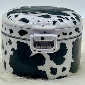 May include: A round, white container with a black cow print design. The container has a white lid and a white latch closure. The container is sitting on a white, fluffy surface.