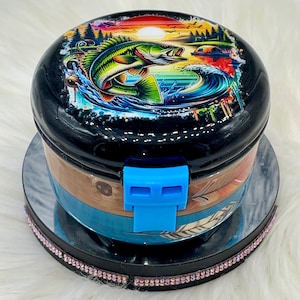 May include: A black fishing tackle box with a colorful graphic of a leaping fish, waves, and a sunset. The box has a blue clasp and a wood-grain and feather design on the sides. The box is on a mirrored surface with a pink rhinestone border.