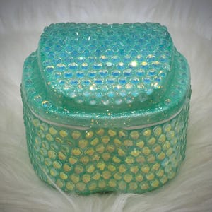 May include: A small, square-shaped container with a lid, fully covered in iridescent, light mint green rhinestones. The surface features a dense, uniform pattern of faceted crystals that reflect light, creating a shimmering effect across the entire exterior.