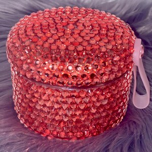May include: A pink container with a lid covered in pink rhinestones. The container has a pink handle.