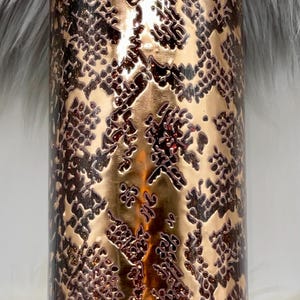 May include: A cylindrical tumbler with a metallic rose gold finish featuring a dark brown snakeskin pattern. The surface has a glossy, smooth texture with embedded glitter accents throughout the design and a clear plastic lid on top.