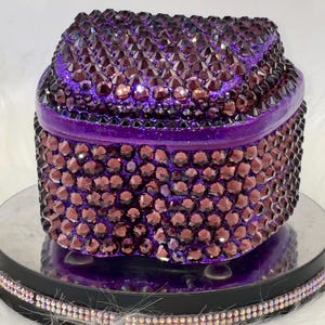 May include: A small, rounded storage box completely covered in deep purple, faceted crystals. The lid and the main body of the container are densely encrusted with these reflective, dark purple stones, creating a uniform, textured, and sparkling appearance.
