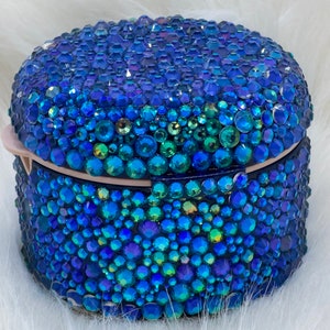 May include: A bedazzled, oval-shaped box covered in blue and teal rhinestones. The rhinestones vary in size and shape, creating a textured surface. The box has a hinged lid and a light pink clasp. The overall effect is a sparkling, decorative item.
