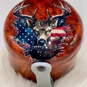 May include: A round, wooden-look badge reel featuring a deer head with large antlers, superimposed over a distressed American flag design. The reel has a white plastic clip and a retractable cord. The overall color scheme is brown, red, white, and blue.