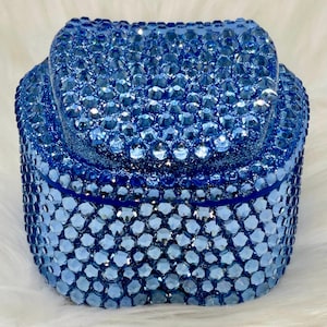 May include: A small, rectangular storage box completely covered in faceted, light blue and dark blue crystals. The lid features a slightly raised, rounded top section, and the entire exterior surface is densely encrusted with shimmering blue stones.
