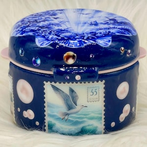 May include: A round, blue ceramic trinket box with a lid featuring a landscape scene. The box has a pink rim and handle, and is decorated with a postage stamp design of a seagull over water. The box is adorned with white and pink accents.