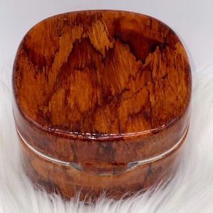 May include: A small, oval-shaped wooden box with a hinged lid. The box is made of a rich, reddish-brown wood with a unique grain pattern. A thin silver band encircles the box at the midsection, and the box is resting on a white, fluffy surface.