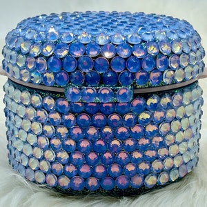 May include: A small, round, blue container covered in iridescent rhinestones. The container has a hinged lid and is perfect for storing small items.