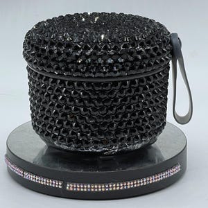 May include: A black, bedazzled container with a matching lid and a gray handle. The container is covered in small, black rhinestones. The base is black and has a band of iridescent rhinestones around the edge.