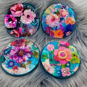 May include: Four round coasters with a turquoise border and colorful floral designs. Each coaster features a different floral pattern with vibrant colors and abstract details.