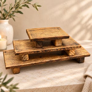 May include: A set of three rectangular wooden risers with a dark brown finish and visible wood grain. Each riser features a flat top surface supported by four short, block-style legs, arranged in a tiered stack from smallest to largest.