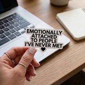 May include: A white die-cut sticker with a black border and black text that reads "EMOTIONALLY ATTACHED TO PEOPLE I'VE NEVER MET" with a small black heart icon centered at the bottom of the text. The sticker has a matte finish and clean edges.