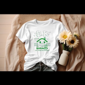 May include: White t-shirt with a green graphic featuring the text "DK" and a house design. Below the house is the text "NANAbnb Let's hang out together!" The shirt is knotted at the waist and paired with blue jeans. A small vase with flowers is to the right.