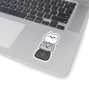 May include: A sticker featuring three stacked cartoon cats. The cats are grey, light grey, and white. The cats have black eyes and black outlines.
