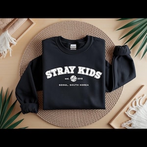 May include: Black crew neck sweatshirt featuring the white text "STRAY KIDS". Below the text are the words "est. 2018" and "SEOUL, SOUTH KOREA". The sweatshirt is presented on a woven mat with green foliage.